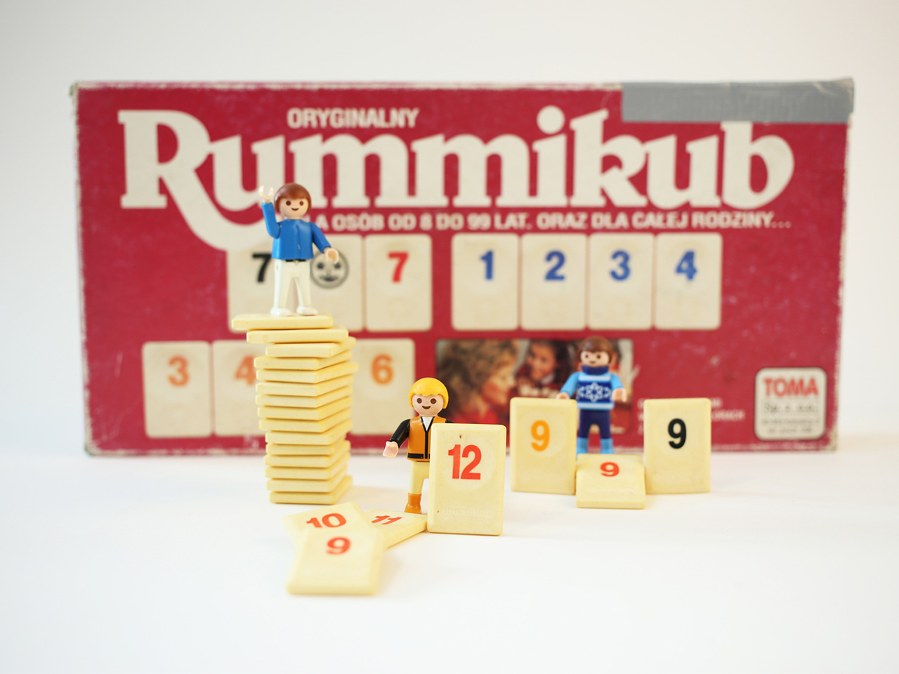 Zdjęcie, na którym jest pudełko z grą Rummikub, małe ludziki i kamienie do gry z cyframi